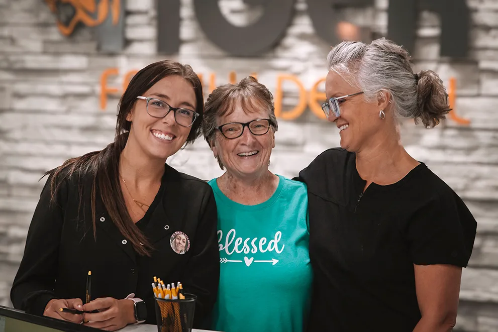 Three women smiling and standing together in a dental office reception area. The woman in the middle, wearing a turquoise shirt with the word Blessed printed on it, appears to be a happy dental patient. She is flanked by two dental staff members dressed in professional attire, both wearing glasses and smiling warmly. The setting includes a modern stone wall with a partially visible logo for a dental clinic in the background, and friendly environment. The image conveys warmth, patient satisfaction, and a strong sense of community care in a family dental practice.