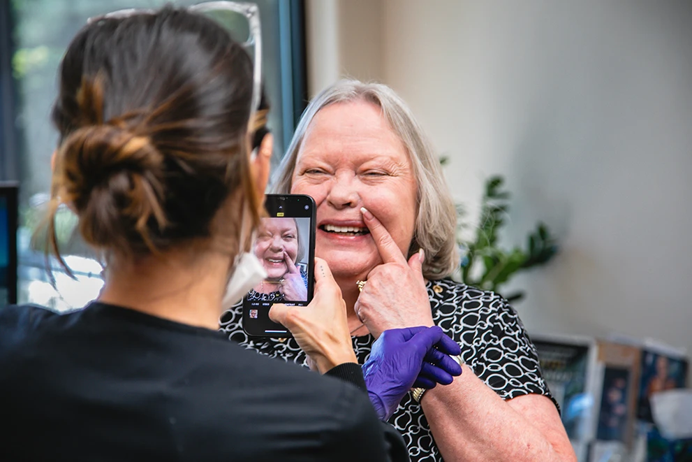 A smiling elderly woman at a dental clinic points to her teeth while a dental professional takes a photo of her using a smartphone. The woman appears happy and is likely showing off recent dental work, such as new dentures or dental implants. The dental professional wears gloves and a mask, suggesting a clinical setting. The smartphone screen shows a clear image of the woman’s joyful expression, capturing the moment. The background includes dental office decor and indoor plants, enhancing the professional yet welcoming environment.